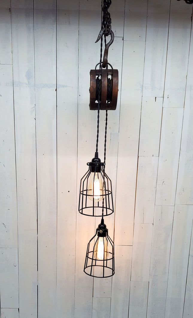 Two offset 5 1/2-inch diameter cage lamp shades housing Edison bulbs, suspended from a double wheel barn pulley, this pendant light hangs 36 inches, and has a 24-inch hanging chain. Lights are Illuminated.