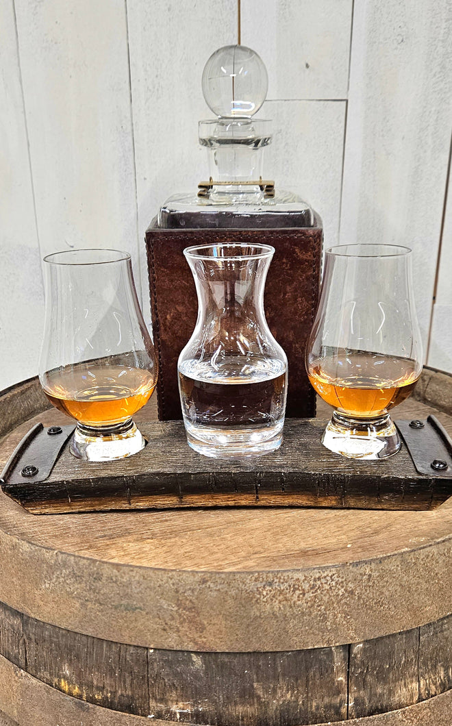 A whiskey barrel stave coaster sporting 3 indents, with a Glencairn glass at each end, and a small carafe in the middle.  The coaster sits on a barrel top. A whiskey decanter sits behind the coaster and the glasses hold whiskey and water.
