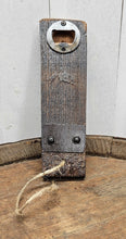 Load image into Gallery viewer, A barrel stave bottle opener with indent cap remover and a barrel ring accent, sits on top of a whiskey barrel. The opener has a jute rope cord at the bottom .
