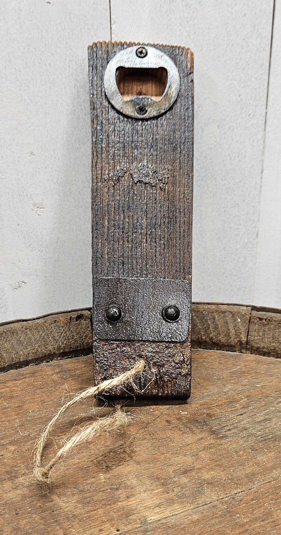 A barrel stave bottle opener with  indent cap remover and a barrel ring accent, sits on top of a whiskey barrel.  The opener has a jute rope cord at the bottom .
