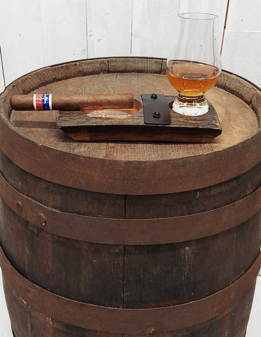 Showing a cigar and whiskey glass, this genuine whiskey barrel stave, accented with a rustic barrel ring with a felt bottomed Glencairn Glass indent, and a copper bottomed cigar receptacle, measures 8” L x 4” W. 