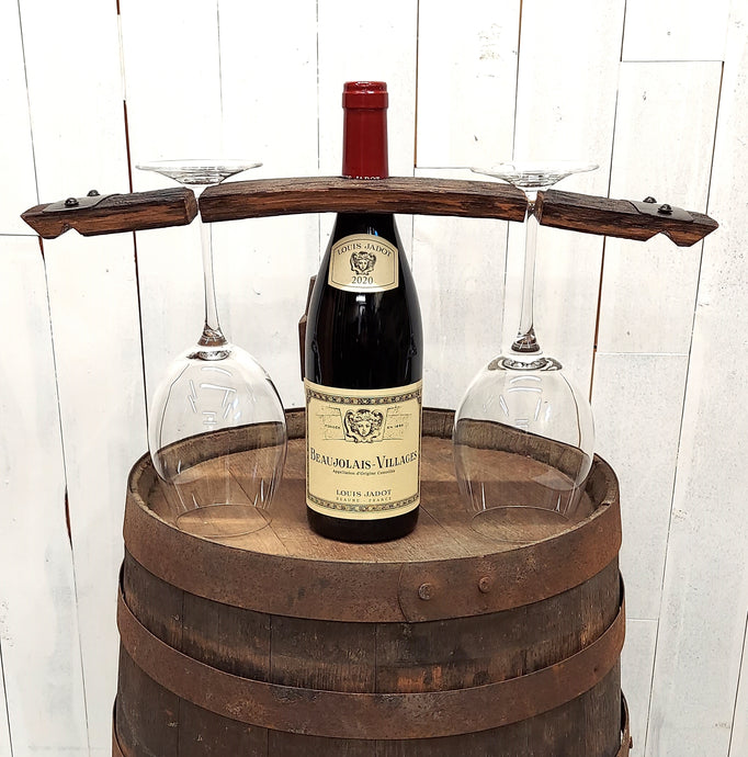 Wine bottle centered on a wooden barrel top, with a rustic barrel stave set over the bottle neck, and a stemmed wine glass hanging  on each side of the bottle.